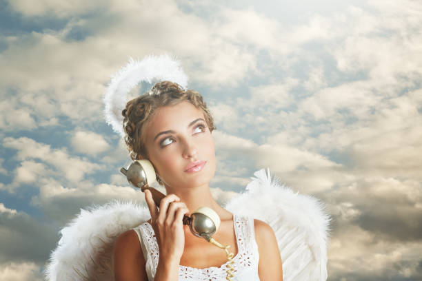 An angel in despair holds her head from what is happening in the world. A blonde in white clothes and with big white angel wings touches her hair with both hands under a clear blue sky..