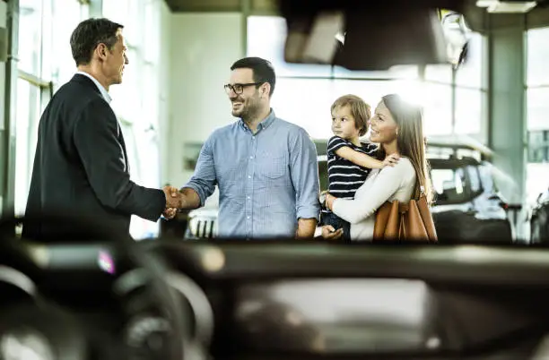 Happy family came to an agreement with salesperson in a car showroom. The view is through windscreen. Men are shaking hands.