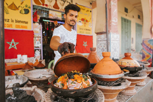 Young seller cooking vegetable tajines in Morocco. Rissani, Morocco - September 18th, 2019: Young moroccan man cooking tajine with vegetables in the street, and showing it to the tourist to sell meal, in Rissani, Morocco. tajine stock pictures, royalty-free photos & images