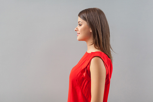 Profile side view portrait of happy beautiful brunette young woman in red shirt standing and looking forward with toothy smile and happiness. indoor, studio shot, isolated on gray background.