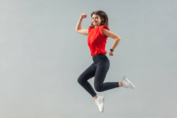 pleine longueur de la jeune femme belle heureuse de brunette dans la chemise rouge occasionnelle sautant vers le haut, sourire denté et regardant l'appareil-photo avec le bonheur - lenght photos et images de collection