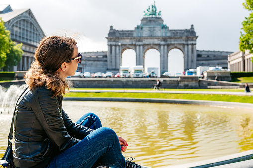 Young Caucasian woman sitting on edge of fountain on the arcade du Cinquantenaire and the north hall in the Cinquantenaire park in Brussels, Belgium.