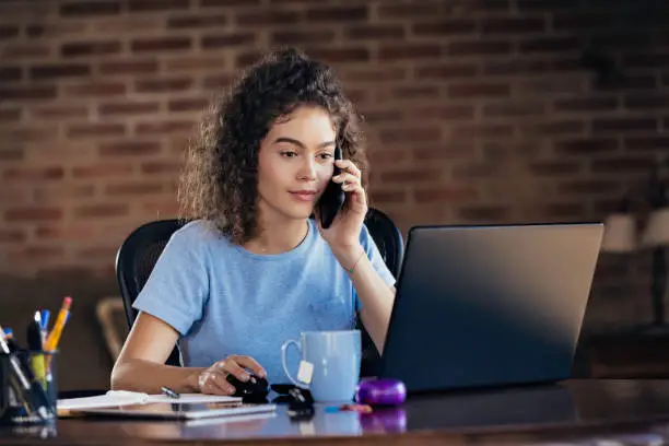 Photo of Multi-tasking Hispanic curly hair young woman using laptop and using mobile phone working at home office