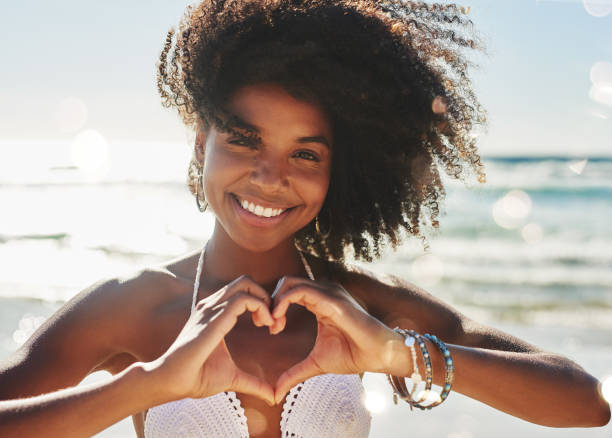 Sun and sea sounds like an awesome summer to me Portrait of a beautiful young woman making a heart shaped gesture with her hands at the beach black woman bathing suit stock pictures, royalty-free photos & images