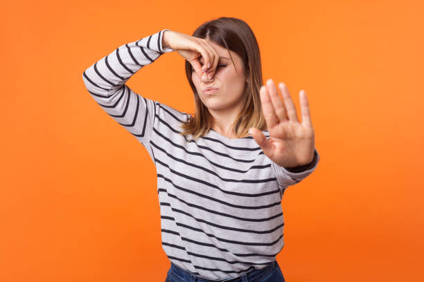 schlechter geruch. porträt der jungen frau mit braunen haaren in langärmeligem gestreiftem hemd. indoor-studio-aufnahme isoliert auf orangefarbenem hintergrund - facial expression unpleasant smell shirt caucasian stock-fotos und bilder