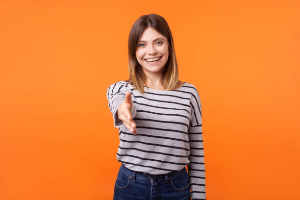 retrato da mulher nova amigável com cabelo marrom na camisa listrada da luva longa. estúdio interno disparado isolado no fundo alaranjado - isolated smiling business person handshake - fotografias e filmes do acervo