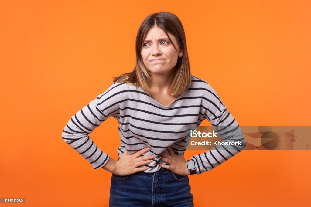 Portrait of unhappy ill young woman with brown hair in long sleeve striped shirt. indoor studio shot isolated on orange background Portrait of unhappy ill woman with brown hair in long sleeve striped shirt standing, holding her belly with hands, stomach cramps or period pain. indoor studio shot isolated on orange background Women Stock Photo