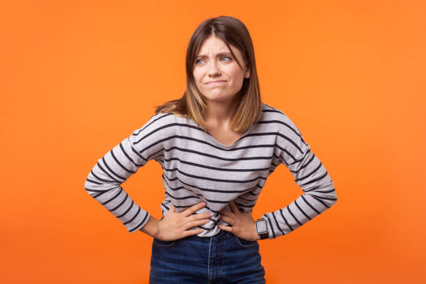 Portrait of unhappy ill young woman with brown hair in long sleeve striped shirt. indoor studio shot isolated on orange background Portrait of unhappy ill woman with brown hair in long sleeve striped shirt standing, holding her belly with hands, stomach cramps or period pain. indoor studio shot isolated on orange background pms stock pictures, royalty-free photos & images