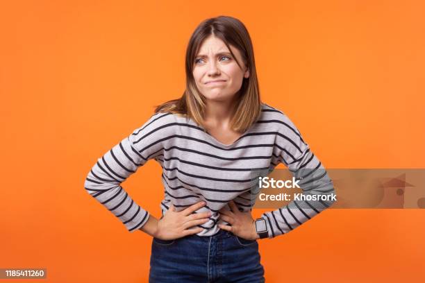 Portrait Of Unhappy Ill Young Woman With Brown Hair In Long Sleeve Striped Shirt Indoor Studio Shot Isolated On Orange Background Stock Photo - Download Image Now