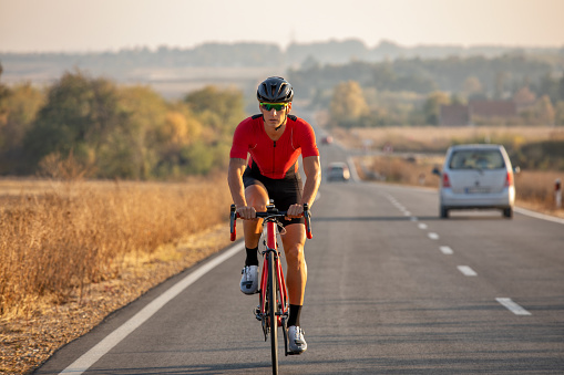 Young professional athlete riding bicycle for exercise on the road through the countryside. He is well equipped, with protective helmet, sunglasses, black and red jersey. Photo taken when cyclist is in motion, from car in motion, for speed effect.