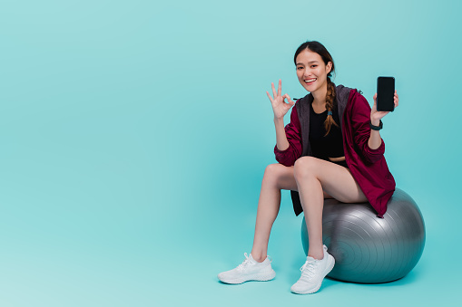 Asian beautiful happy woman holding smartphone and sitting on fit ball after exercise isolated on blue color background wite studio shot.Concept of slim and healthy girl workout.