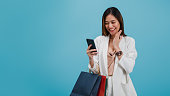 アジアの美しい女性ブロガーは、コピースペースと青い色の背景に隔離されたショッピングバッグでオンラインスマートフォンショッピングを使用しています。プロモーションと販売による�
