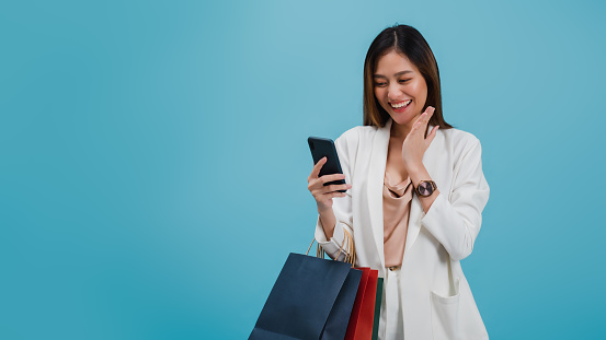 Asian beautiful women blogger are using smart phone shopping online with a shopping bag isolated in blue color background with copy space.Concept of online shopping business with Promotion and Sale.