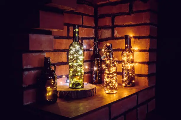 Photo of Lot of different vintage style beer bottles and green wine bottles decorated with wire string micro led lights on shelf, red brick wall background. Illuminated at night.