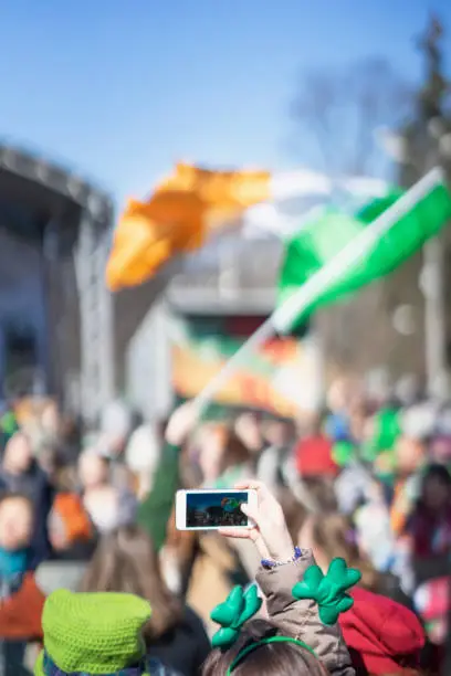 Female hands with mobile phone, making photo of traditional national Irish holiday party, carnival of St. Patrick's Day on a smartphone, selective focus. Vertical background