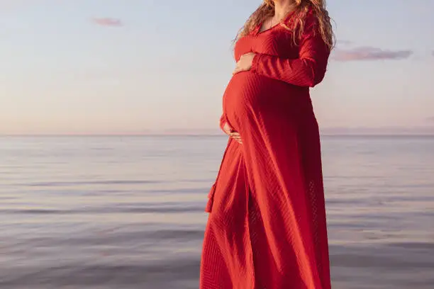 Portrait of beautiful pregnant woman in a red dress touching her stomach, standing outside by the ocean in beautiful autumn evening sunlight, stock picture.