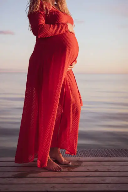 Portrait of beautiful pregnant woman in a red dress touching her stomach, standing outside by the ocean in beautiful autumn evening sunlight, stock picture.