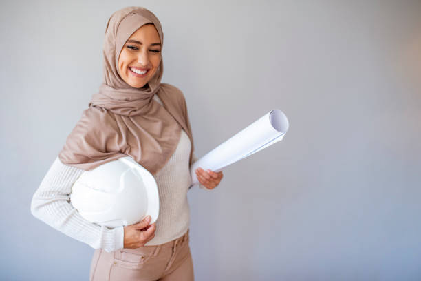 Ingenieur Arabic muslim woman with hijab holding protective helmet. Young Muslim architect-woman wearing a protective helmet standing on the grey background. Pretty arabic architect woman isolated shot in studio. Ingenieur Arabic muslim woman with hijab holding protective helmet. ingenieur stock pictures, royalty-free photos & images