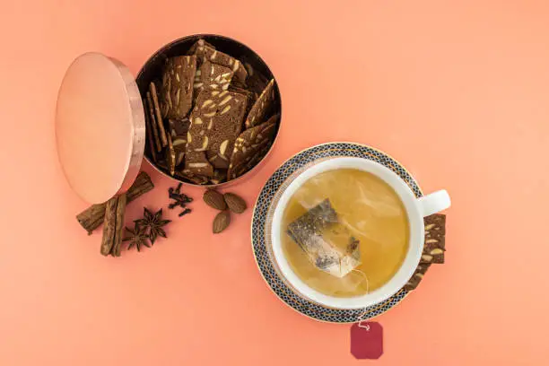 Studio image of a steaming teacup with a bronze tin with Danish brunkager, and some of the ingredients of the tea and of the cakes on the table. The background is living coral colored (PANTONE 2019 year color)