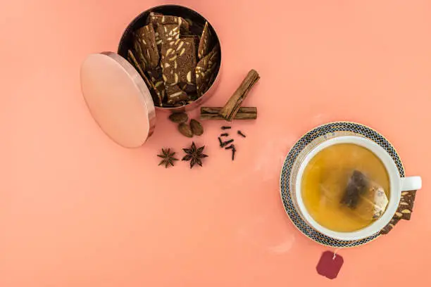 Studio image of a steaming teacup with a bronze tin with Danish brunkager, and some of the ingredients of the tea and of the cakes on the table.

The background is living coral colored  (PANTONE 2019 year color)
