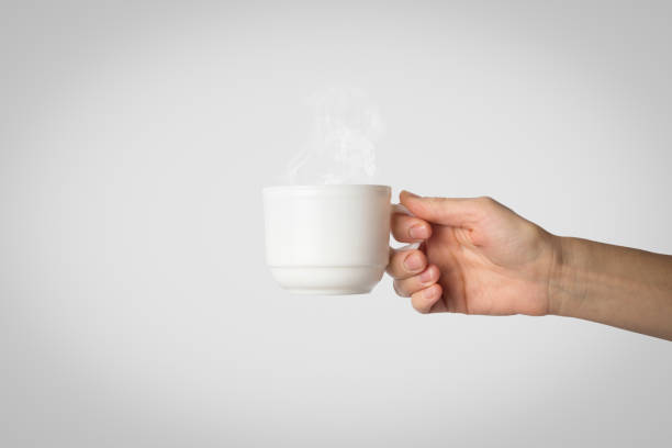 istock Female hand holding a white cup with hot coffee on a gray background. Side view 1185435504