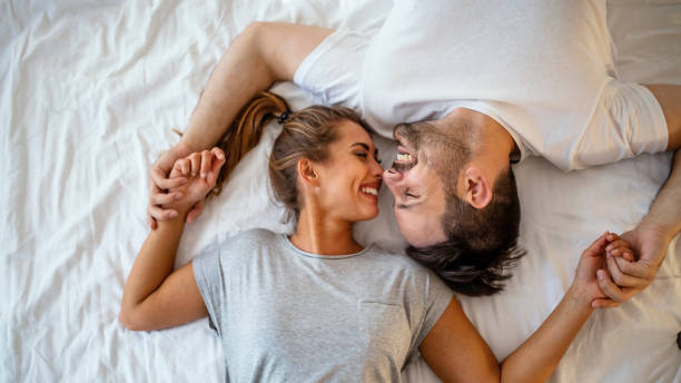 Beautiful pair of lovers hug and kiss Guy and a girl in a cozy home environment. Happy man and woman lying in the bedroom stock photo. Top view of smiling young couple cuddling in bed in morning.  Beautiful pair of lovers hug and kiss young couple stock pictures, royalty-free photos & images