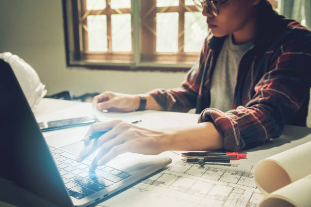 istock Engineering Asian man architects wear glasses  holding pencil working On laptop with phone,hat and blueprint Behind the sun shining through the window. 1185455189
