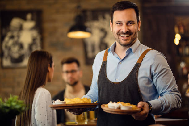 Happy waiter holding plates with food and looking at camera in a pub. Portrait of happy waiter working in a pub and serving food while looking at camera. waiter stock pictures, royalty-free photos & images