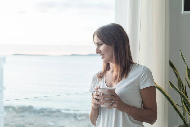 istock Adult woman standing while looking through window tasting coffee 1185492571