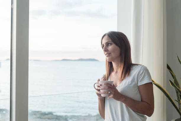 istock Adult woman standing while looking through window tasting coffee 1185505654