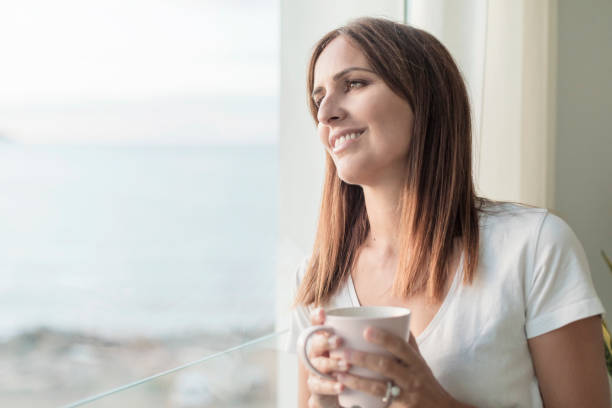 istock Adult woman standing while looking through window tasting coffee 1185505914