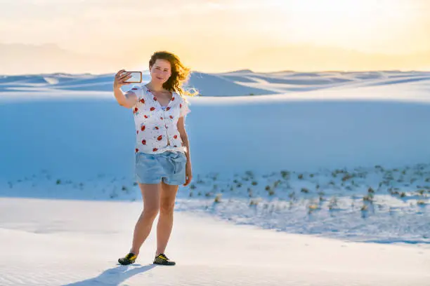 Woman taking selfie picture on sand hill in white sands dunes national monument in New Mexico using phone with wind