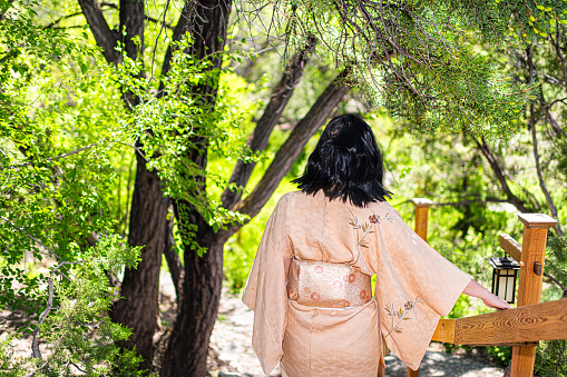 Young woman in kimono costume walking down back holding on wooden railing fence in outdoor garden in Japan with nature view