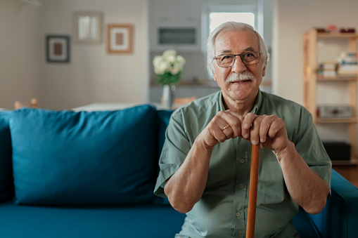 Portrait of happy senior man smiling at home while holding walking cane. Old man relaxing on sofa and looking at camera. Portrait of elderly man enjoying retirement.\