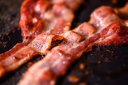 Extreme close up shots of pork bacon sliced and being fried on a black hot pan, under oil. Illustration for meat, eating, meal, breakfast with no people