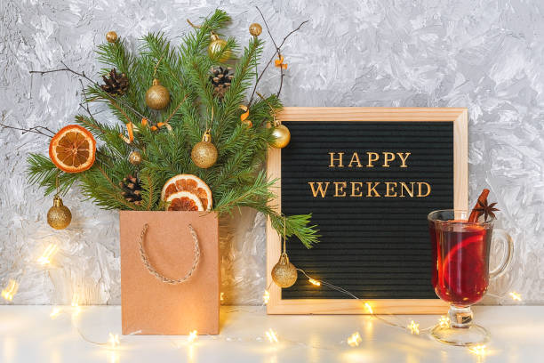 istock Happy Weekend text on black letter board, festive decored christmas tree in craft package, light lamp garland and glass of hot mulled wine with spices on table. Concept Hello winter weekend 1185558458