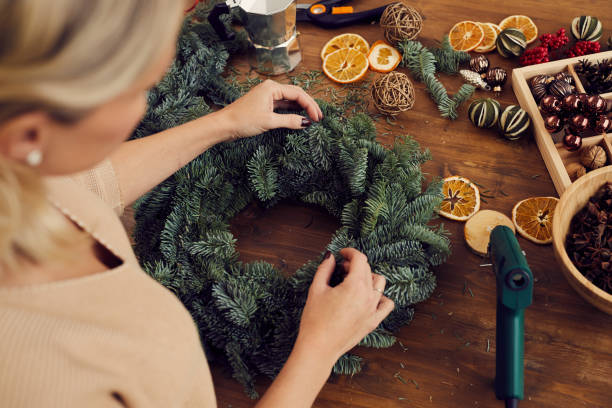 Above view of craftswoman standing at wooden table full of decorations and creating Christmas wreath in workshop Close-up of unrecognizable woman standing at wooden table and using secateurs while cutting fir twig for Christmas wreath christmas human hand christmas ornament decoration stock pictures, royalty-free photos & images