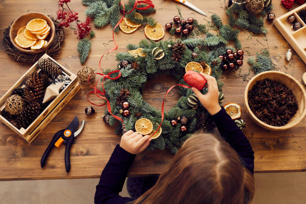hochwinkelansicht der beschäftigten frau, die am schreibtisch steht und dekoratives band verwendet, während sie weihnachtskranz machen - vorbereitung stock-fotos und bilder