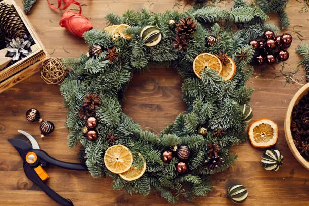 Photo of High angle view of beautiful holiday wreath decorated orange slices, fir tree cones and small balls placed on wooden table among decorations and tools