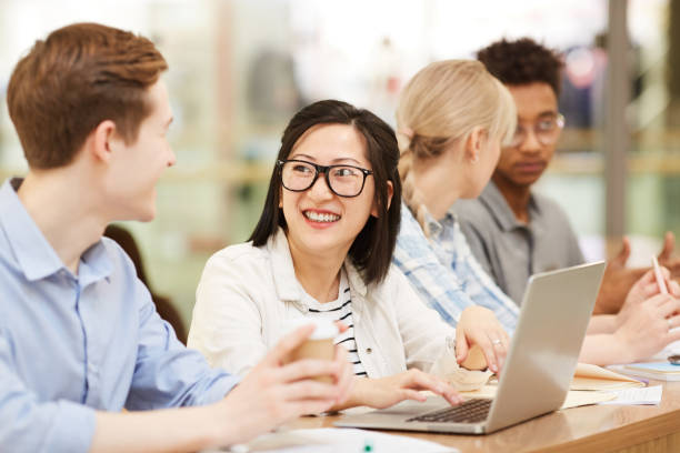 istock Group of cheerful young interracial students sitting at desk in library and discussing ideas while working on educational projects 1185581904