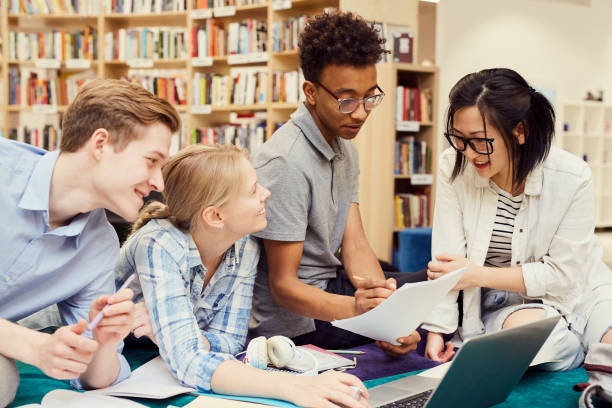 grupa pozytywnych młodych wieloetnicznych studentów siedzących w nowoczesnej bibliotece i oglądania programu nauczania podczas przygotowań do zajęć - multi function zdjęcia i obrazy z banku zdjęć