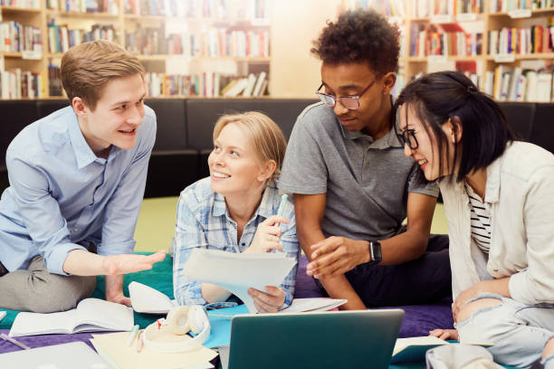 istock Group of cheerful enterprising multi-ethnic students sitting in university library and discussing tasks while preparing for exam together 1185581970