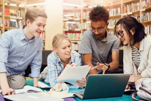 gruppe positiver kreativer multi-ethno-studenten in lässigen outfits, die in der universitätsbibliothek sitzen und beispielarbeit zur vorbereitung auf die projektpräsentation ansehen - multi ethnic group reading stock-fotos und bilder