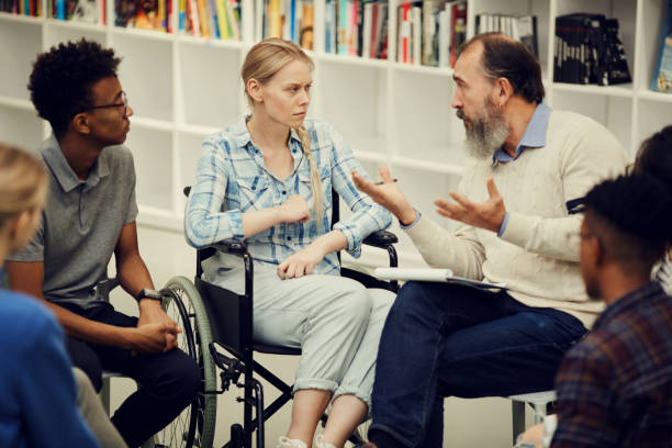 istock Senior bearded therapist sitting in circle of young people and gesturing hands while talking to disabled girl in wheelchair at group therapy 1185582062