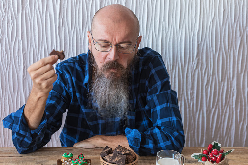 Casual dressed man with long beard sitting at home, eating home made dark chocolate with Christmas cookies, real people