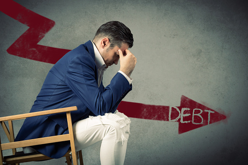 Stressed businessman with headache sitting in front of a wall with debt sign on arrow symbol.