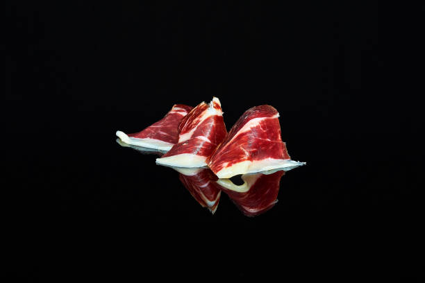 foto de estudio de una serie de rebanadas de jamón 100% ibérico de bellota plegado sobre fondo negro con reflejo. - península ibérica fotografías e imágenes de stock