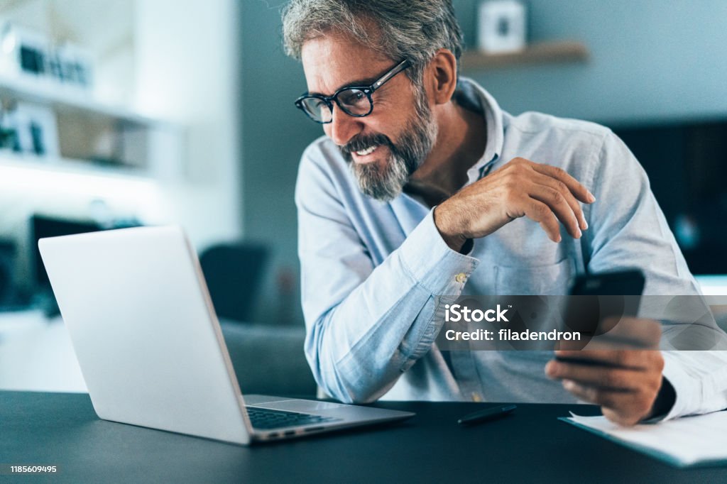 Working at home Mature businessman working at home using lap top and smart phone Laptop Stock Photo