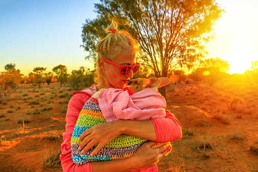 Tourist woman holding ed kissing kangaroo joey at sunset light in Australian outback. Interacting with cute kangaroo orphan. Australian Marsupial in Northern Territory, Central Australia or Red Center