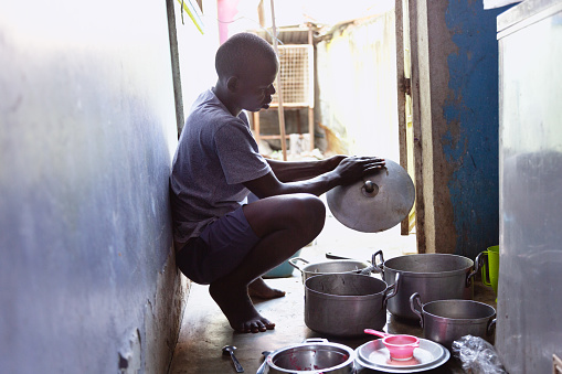Young african man, 23 years old, wearing a  grey shirt and blue shorts, scrubbing pots in a small kitchen with concrete floor. Photo was taken in Dar es Salaam, Tanzania.